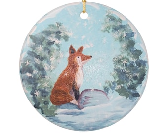 Peut inclure: Une décoration ronde en céramique représentant une scène peinte d'un renard roux assis dans un paysage enneigé. Le renard est entouré de deux sapins vert foncé avec des flocons de neige blancs sur un ciel bleu clair. Un cordon doré est fixé.