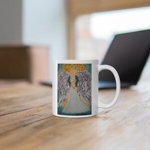 Puede incluir: Taza de cer&aacute;mica blanca con una ilustraci&oacute;n colorida de un &aacute;ngel con alas y un arpa. El &aacute;ngel lleva un vestido azul y tiene el pelo rubio.
