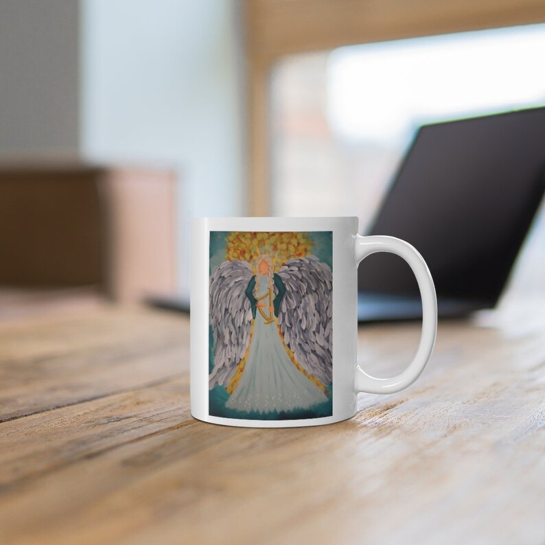Puede incluir: Taza de cer&aacute;mica blanca con una ilustraci&oacute;n colorida de un &aacute;ngel con alas y un arpa. El &aacute;ngel lleva un vestido azul y tiene el pelo rubio.