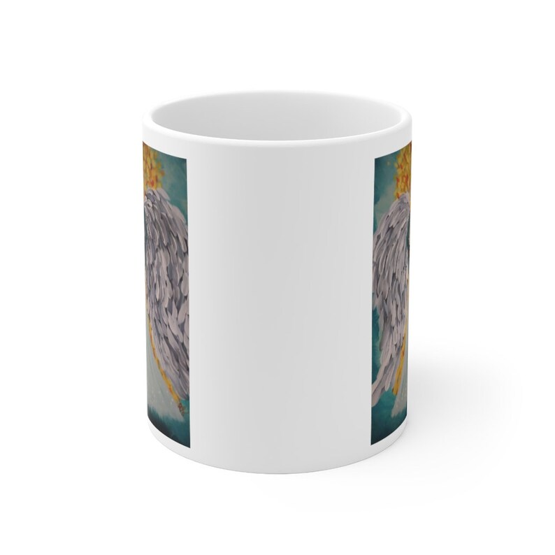 Puede incluir: Taza de cer&aacute;mica blanca con una imagen colorida de alas de &aacute;ngel en ambos lados. Las alas est&aacute;n pintadas en tonos de gris, azul y amarillo.