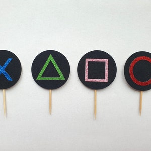 Può includere: Quattro topper per torta circolari neri con diversi simboli colorati. I simboli sono una X blu, un triangolo verde, un quadrato rosa e un cerchio rosso.