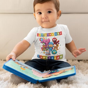 May include: A white t-shirt with the text "BIRTHDAY BOY" and the name "SEBASTIAN" in colorful block letters. The shirt features cartoon characters and a birthday cake. The child is wearing blue jeans and holding a book.