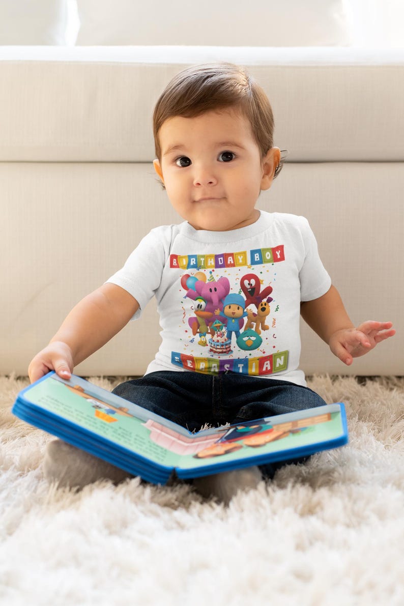 May include: A white t-shirt with the text "BIRTHDAY BOY" and the name "SEBASTIAN" in colorful block letters. The shirt features cartoon characters and a birthday cake. The child is wearing blue jeans and holding a book.