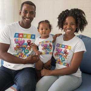 May include: Three people wearing matching white t-shirts and a onesie with a colorful cartoon design. The shirts say "Daddy" and "Mommy" in a block letter style. The baby is wearing a matching onesie. The family is sitting on a blue couch.