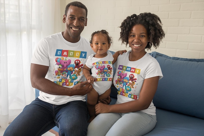 May include: Three people wearing matching white t-shirts and a onesie with a colorful cartoon design. The shirts say "Daddy" and "Mommy" in a block letter style. The baby is wearing a matching onesie. The family is sitting on a blue couch.