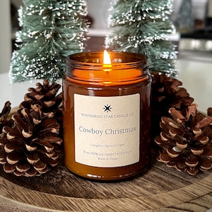 May include: A lit amber glass candle labeled "Cowboy Christmas" sits on a wooden tray surrounded by pine cones and small decorative Christmas trees. The candle's label lists "Campfire | Spice | Juniper" as scents.