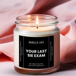 May include: A clear glass candle jar with a black label that reads "Smells Like Your Last Sie Exam". The label also says "No capes required, just a lot of patience". The candle is lit and the flame is visible.