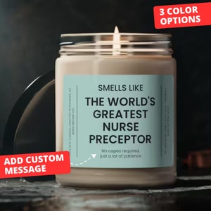 May include: A soy candle with a light blue label that reads "Smells Like The World's Greatest Nurse Preceptor. No capes required, just a lot of patience." The candle is in a clear glass jar with a white lid. The candle is lit and the flame is visible. The background is a dark gray surface.