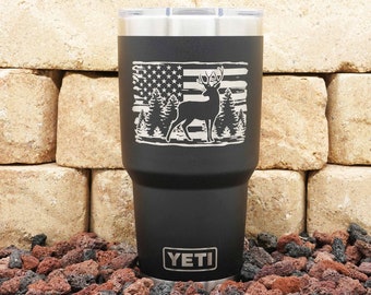 May include: Black Yeti tumbler with a laser engraved design of an American flag and a deer silhouette. The text "YETI" is printed on the bottom of the tumbler.