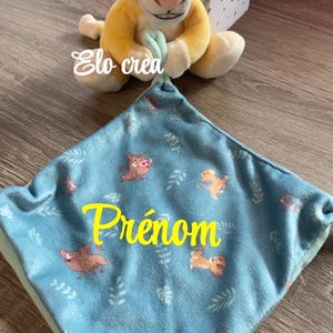 Puede incluir: Un peluche de Simba y una manta azul para bebé. La manta tiene un patrón repetido de animales de dibujos animados y la palabra "Prénom" en amarillo. La manta es azul claro y tiene una textura suave.