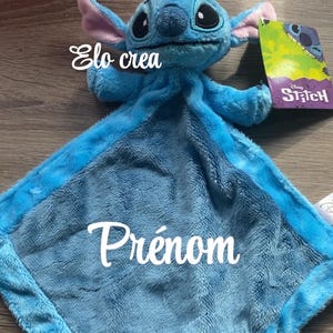 May include: A blue plush toy of the character Stitch from Disney's Lilo & Stitch. The toy has a soft, textured blanket with the word "Pr&eacute;nom" embroidered on it. The tag reads "Stitch". The text "Elo crea" is also visible.