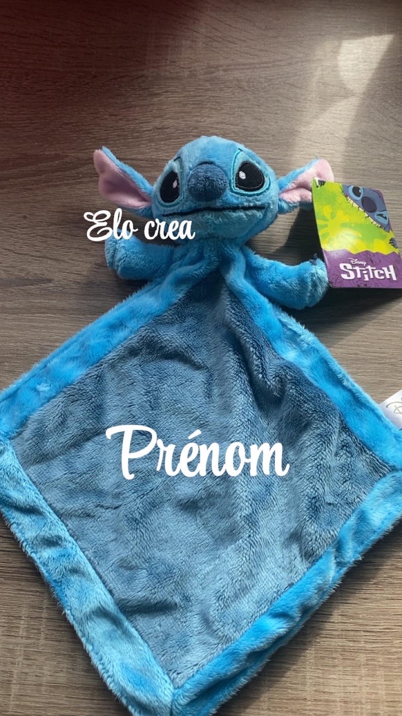 May include: A blue plush toy of the character Stitch from Disney's Lilo & Stitch. The toy has a soft, textured blanket with the word "Pr&eacute;nom" embroidered on it. The tag reads "Stitch". The text "Elo crea" is also visible.