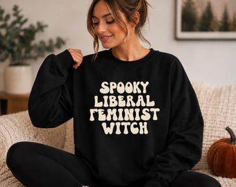 May include: A black long-sleeve crewneck sweatshirt featuring the text SPOOKY LIBERAL FEMINIST WITCH printed in a large, cream-colored, wavy, retro-style font across the center of the chest. The garment has a ribbed neckline and cuffs.