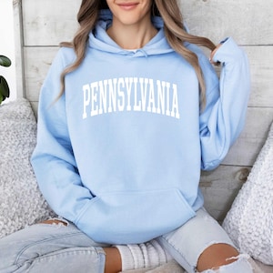 May include: A light blue hoodie with the word "PENNSYLVANIA" printed in white across the chest. The hoodie has a drawstring hood and a front pocket. The person is wearing ripped blue jeans.