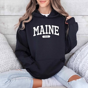 May include: Black hooded sweatshirt with the word "MAINE" in white block letters across the chest, with "USA" underneath. The sweatshirt has a front pocket and a drawstring hood. The person is wearing light blue jeans.