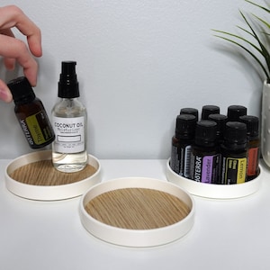 May include: Three white circular trays with wooden inserts, each holding essential oil bottles. The bottles are labeled with names like "Lavender", "Lemon", and "Thyme". One tray also holds a clear bottle labeled "Coconut Oil Moisturizer".