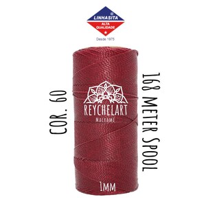 Può includere: Un rotolo di cordoncino rosso da 1 mm di spessore per macramè. Il rotolo è etichettato "168 Meter Spool" e "COR. 60". L'etichetta include anche il nome del marchio "REYCHELART MACRAMÉ".