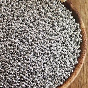 May include: A wooden bowl filled with small, shiny, silver beads. The beads are densely packed, creating a textured surface. The bowl sits on a wooden surface, with dried white flowers in the background. The beads are likely for jewellery making.