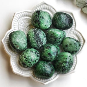 Peut inclure: Un ensemble de pierres vertes polies avec des mouchetures noires, disposées sur une assiette en céramique blanche ornée. Les pierres sont ovales et de tailles variables. Des feuilles d'eucalyptus sont en arrière-plan.