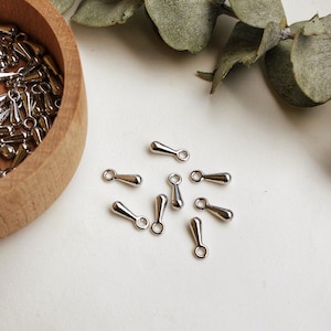 May include: A collection of small, silver-colored metal findings, some scattered on a white surface, others in a wooden bowl. The findings have a teardrop shape with a small loop at one end, suitable for jewelry making.