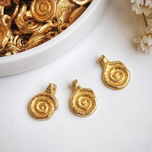 Pode incluir: Três pingentes de metal dourado com um design em espiral. Os pingentes estão sobre uma superfície branca.