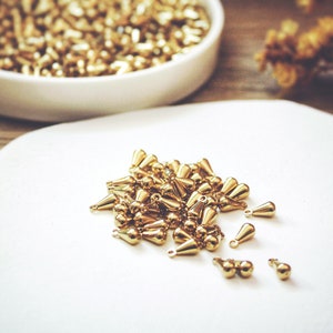 May include: A white plate with a pile of gold teardrop-shaped beads. The beads are scattered around the plate, with some of them overlapping.