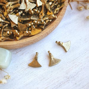 May include: A selection of small, gold-coloured metal axe charms. Some are in a wooden bowl, while three are scattered on a white, textured surface. The axes have a textured finish and a loop at the top for attaching to jewellery.