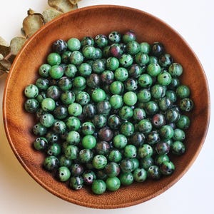 Puede incluir: Un cuenco de madera lleno de cuentas de piedras preciosas verdes y rojo oscuro. Las cuentas son redondas y tienen un aspecto moteado. El cuenco es de madera marrón claro y está sobre una superficie blanca. Hojas secas en el fondo.