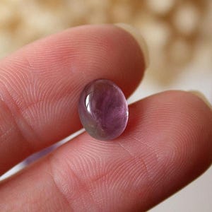 May include: A smooth, oval gemstone in shades of purplish-pink, held between fingers. The cabochon-cut stone displays a translucent quality and internal color variations. This polished gem is likely intended for use in jewellery, with a rounded surface.