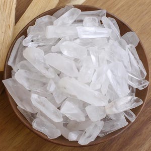 May include: A wooden bowl filled with clear quartz crystal points. The crystals are various sizes and shapes, with a translucent appearance. Two pieces of light-coloured wood are placed to the left of the bowl, on a wooden surface.