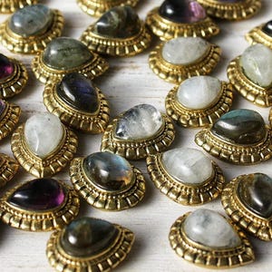 Può includere: Assortimento di cabochon di pietre preziose incastonati in montature in metallo dorato. Le pietre includono ametista, pietra di luna e labradorite.