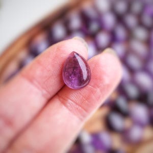 May include: A polished, teardrop-shaped amethyst gemstone held in a hand. The gem is a deep purple colour with internal patterns and a glossy surface. Other similar gemstones are visible in the background, out of focus.