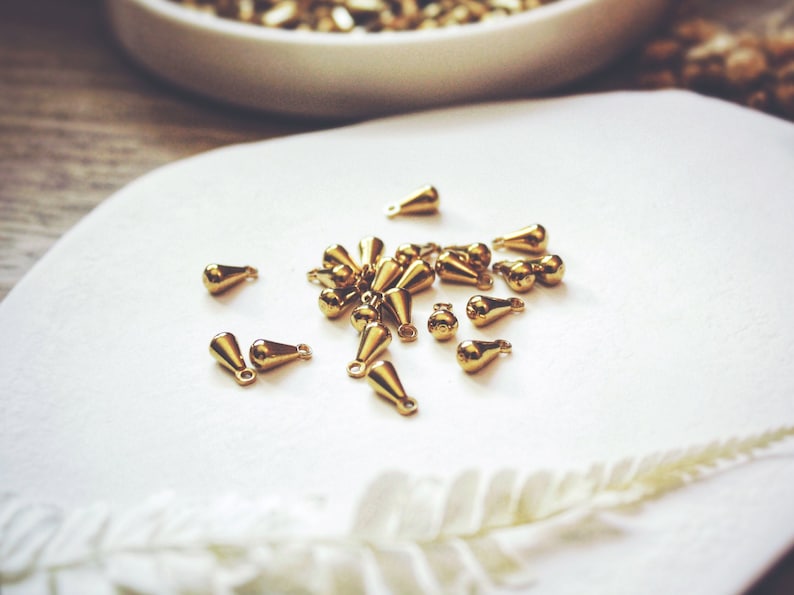 May include: A collection of gold-colored teardrop-shaped metal charms with a loop at the top. The charms are scattered on a white surface.