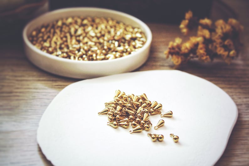 May include: A white plate with a pile of gold teardrop-shaped beads. The beads are scattered around the plate, with some of them overlapping.