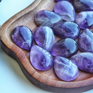 May include: A collection of polished amethyst gemstones in a teardrop shape, displayed in a wooden bowl. The gemstones exhibit a deep purple hue with lighter, translucent areas. The bowl has a natural wood grain.