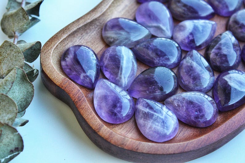 May include: A collection of polished amethyst gemstones in a teardrop shape, displayed in a wooden bowl. The gemstones exhibit a deep purple hue with lighter, translucent areas. The bowl has a natural wood grain.