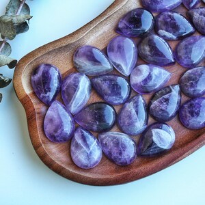 May include: A collection of polished amethyst gemstones in a teardrop shape, displayed on a wooden tray. The gemstones vary in shades of purple, with some showing internal patterns. A sprig of dried leaves is visible in the upper left corner.