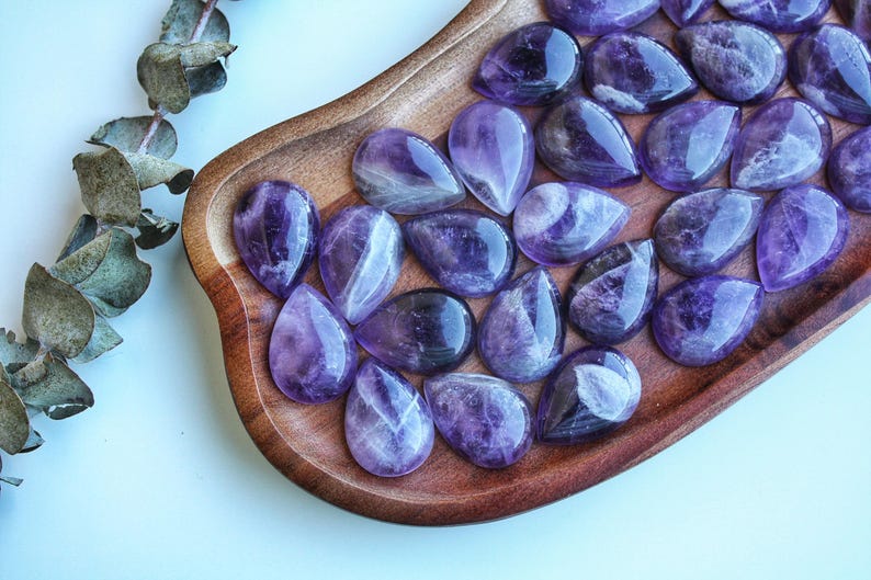 May include: A collection of polished amethyst gemstones in a teardrop shape, displayed on a wooden tray. The gemstones vary in shades of purple, with some showing internal patterns. A sprig of dried leaves is visible in the upper left corner.