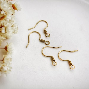 Peut inclure: Quatre crochets de boucles d'oreilles dorés avec un détail de ressort hélicoïdal et un crochet incurvé. Les crochets sont disposés sur une surface blanche, avec des fleurs blanches séchées dans le coin supérieur gauche. Fournitures de fabrication de bijoux.