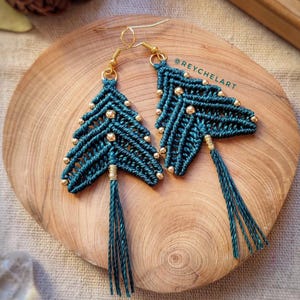 May include: A pair of teal blue macrame earrings with gold beads and tassels. The earrings are in the shape of a triangle with a pointed tip. The earrings are hanging from gold hooks. The earrings are on a wooden surface. @reychelar
