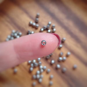 May include: A close-up of a single silver bead, held between a finger and thumb. The bead is round and has a small hole in the centre. There are many other silver beads scattered around the bead, on a wooden surface.