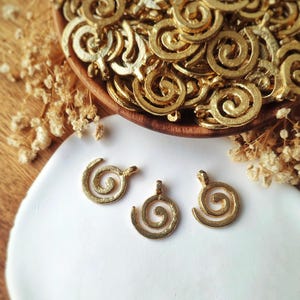 Op de afbeelding: Een verzameling kleine, goudkleurige metalen bedels in de vorm van spiralen. Elke bedel heeft een cirkelvormig spiraalontwerp met een klein lusje aan de bovenkant voor bevestiging. De bedels hebben een getextureerde, matte afwerking.
