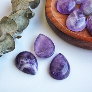 May include: Close-up of polished amethyst gemstones in a teardrop shape. The gemstones are a deep purple color with white veining. Some are displayed in a wooden bowl, while others are scattered on a white surface. A sprig of green leaves is in the background.