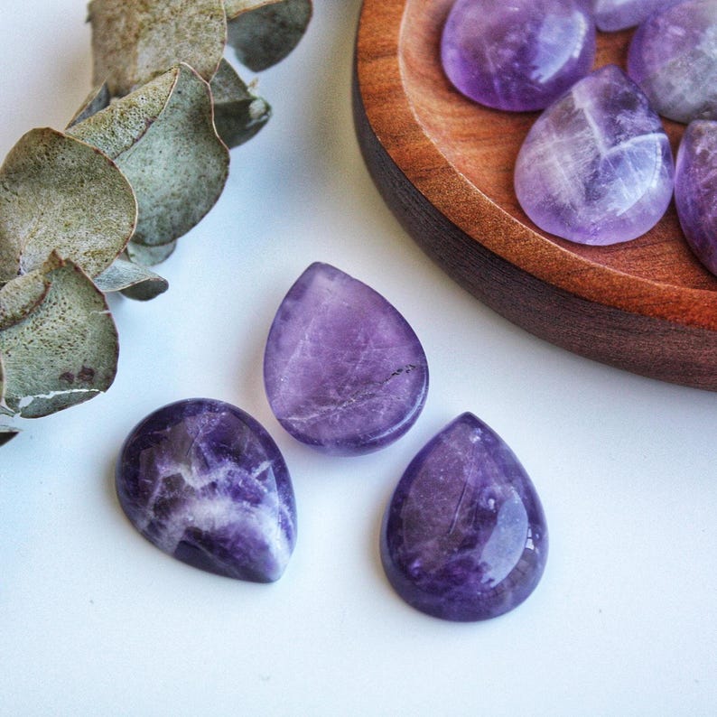 May include: Close-up of polished amethyst gemstones in a teardrop shape. The gemstones are a deep purple color with white veining. Some are displayed in a wooden bowl, while others are scattered on a white surface. A sprig of green leaves is in the background.