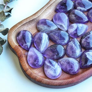 May include: A collection of polished, teardrop-shaped amethyst gemstones displayed on a wooden tray. The gemstones exhibit a range of purple hues, from light lavender to deep violet, with natural patterns. A sprig of dried eucalyptus is in the background.