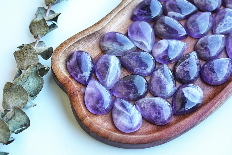 May include: A collection of polished, teardrop-shaped amethyst gemstones displayed on a wooden tray. The gemstones exhibit a range of purple hues, from light lavender to deep violet, with natural patterns. A sprig of dried eucalyptus is in the background.