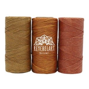 May include: Three spools of macrame cord in shades of brown, orange, and red-brown. The central spool displays the text "REYCHELART MACRAME" in white. The cord is tightly wound around cardboard tubes, highlighting the texture and colour variations.