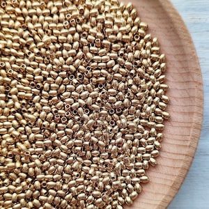 May include: A close-up image of a large quantity of gold-coloured metal beads. The beads are oval-shaped and have a smooth, shiny finish. The beads are arranged in a random pattern, creating a visually appealing texture.