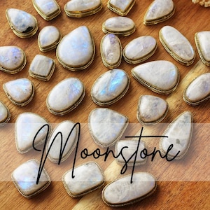Può includere: Una collezione di cabochon di pietra di luna con bordi in metallo dorato. Le pietre sono bianche con sottili riflessi blu e iridescenti. I cabochon hanno varie forme, tra cui ovale, a goccia e quadrata, su una superficie di legno.