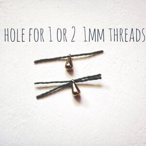 May include: Two gold teardrop-shaped charms with a hole for 1 or 2 mm threads. The charms are hanging from green string.